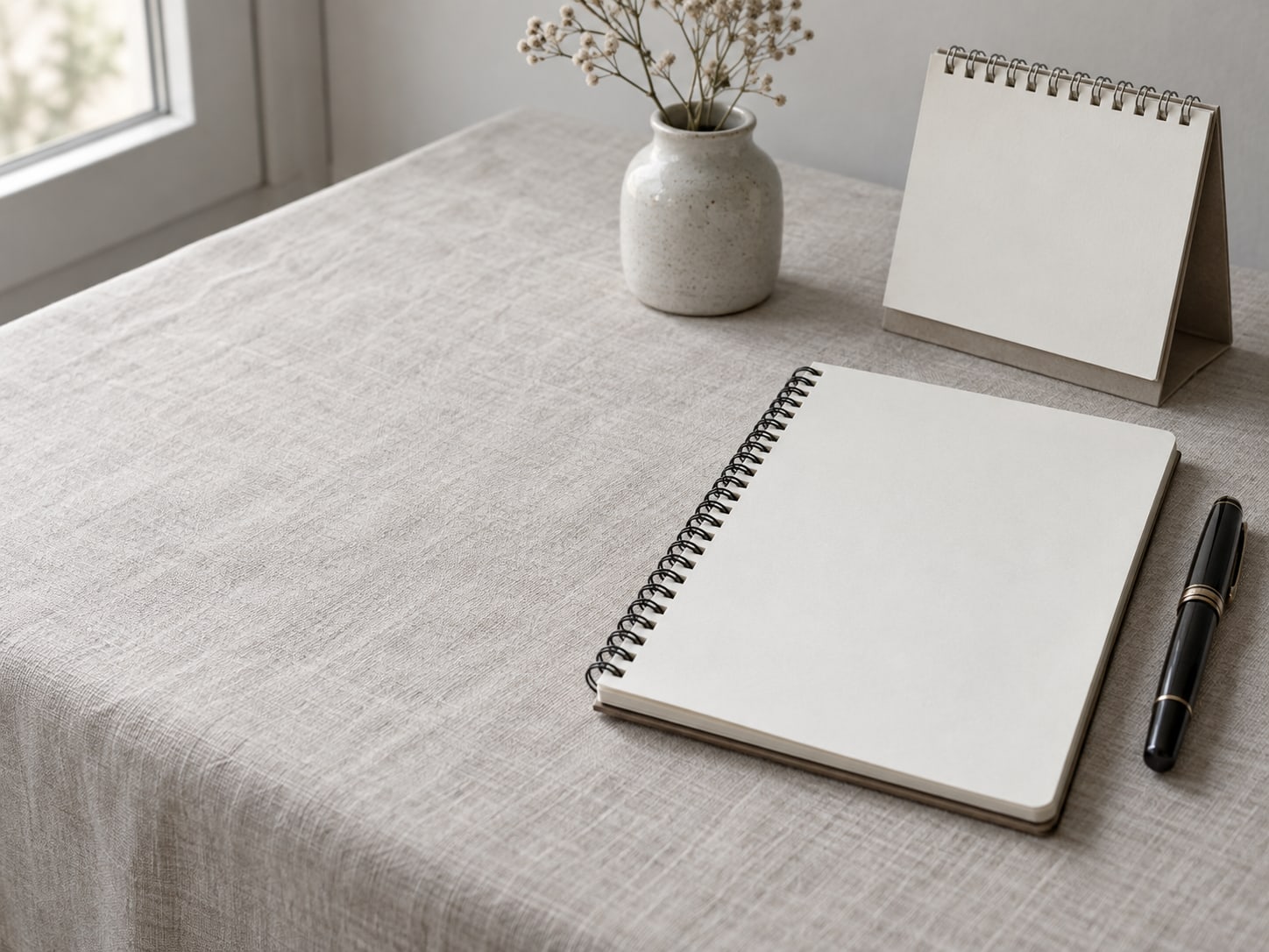 Una libreta espiral abierta en una página en blanco junto a una pluma fuente con tapa y un pequeño calendario de escritorio sin ninguna marca, sobre un mantel de lino pálido.