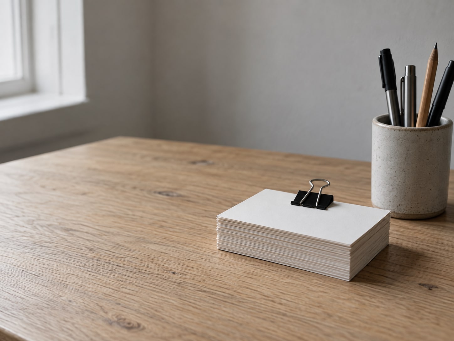 Una pila ordenada de tarjetas en blanco sujeta con un clip de carpeta junto a una pequeña taza de cerámica con plumas, sobre una mesa de roble claro.