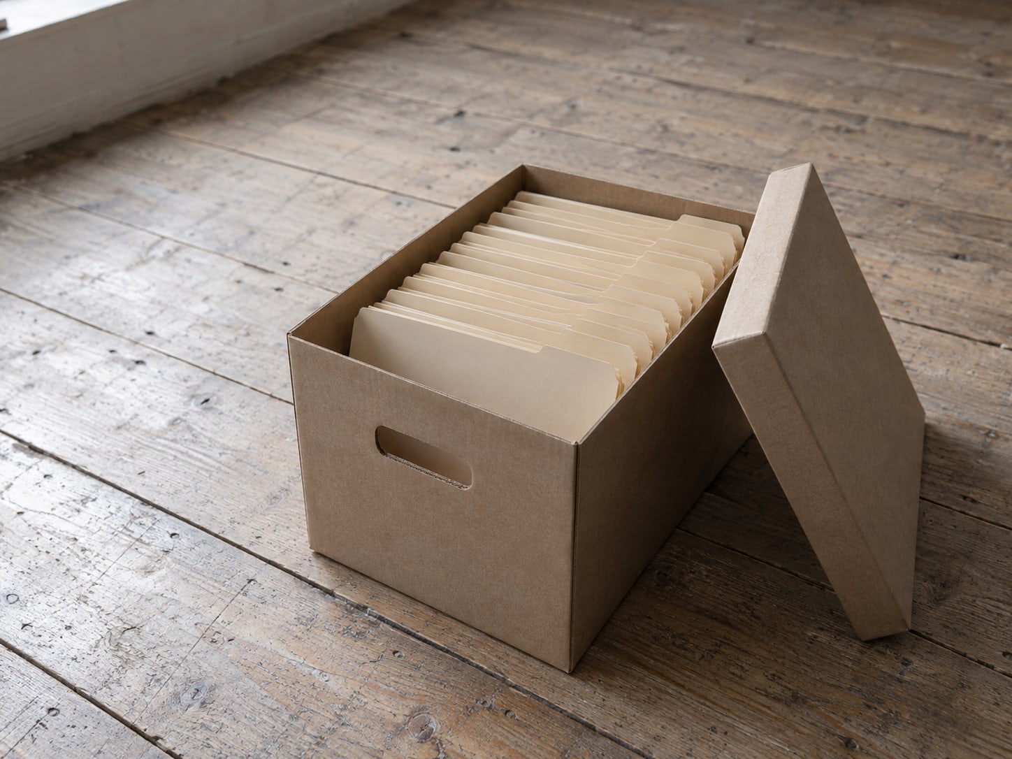 Una caja de archivo de cartón abierta en un piso de madera, con una fila de carpetas manila en blanco, y su tapa recargada al lado.