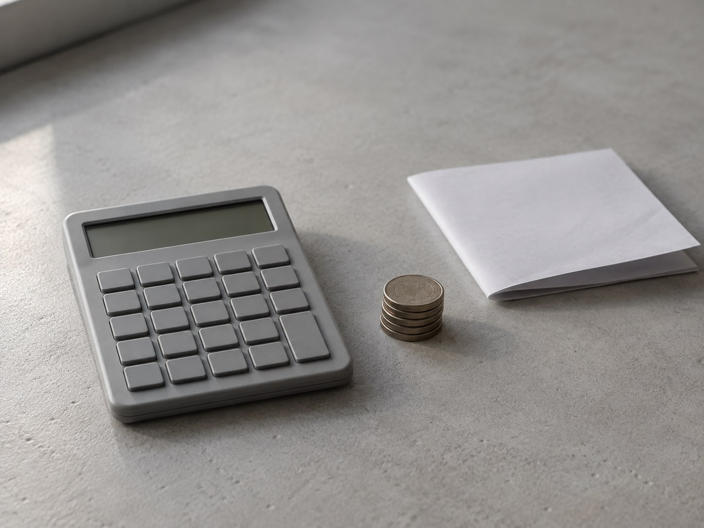 Una calculadora gris sencilla con pantalla en blanco junto a una pequeña pila de monedas y una hoja de papel en blanco doblada sobre un escritorio.