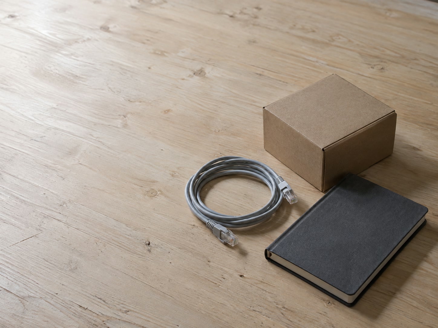 Una pequeña caja de cartón con las solapas cerradas junto a un cable de red enrollado y un cuaderno cerrado sobre una mesa de trabajo limpia.