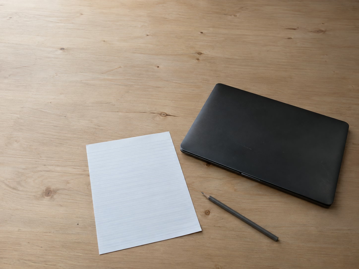 Una laptop cerrada junto a una sola hoja de papel rayado en blanco y un lápiz sobre un escritorio de madera liso.
