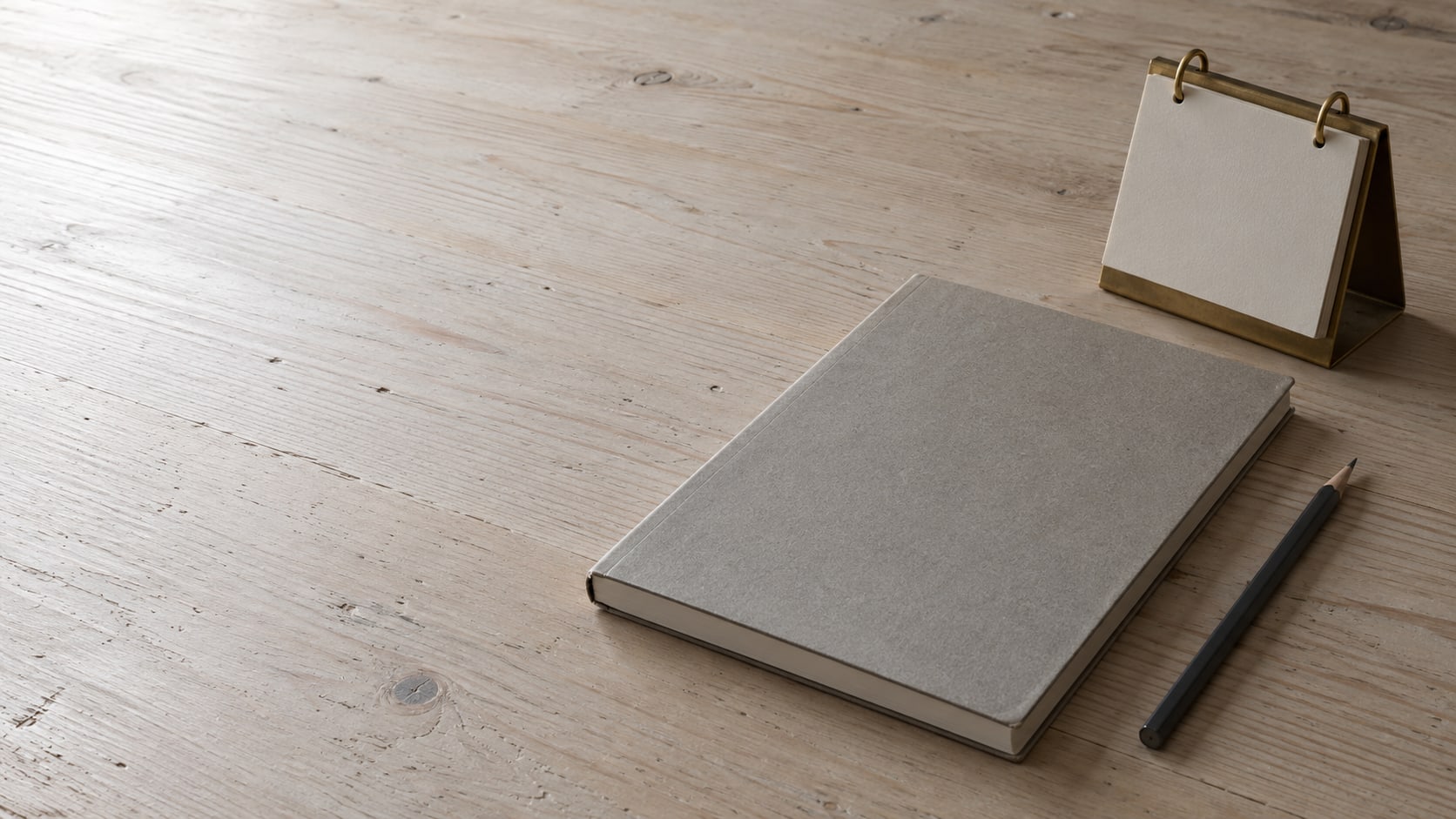 A closed notebook, a pencil and a small brass desk calendar on a pale wooden desk.