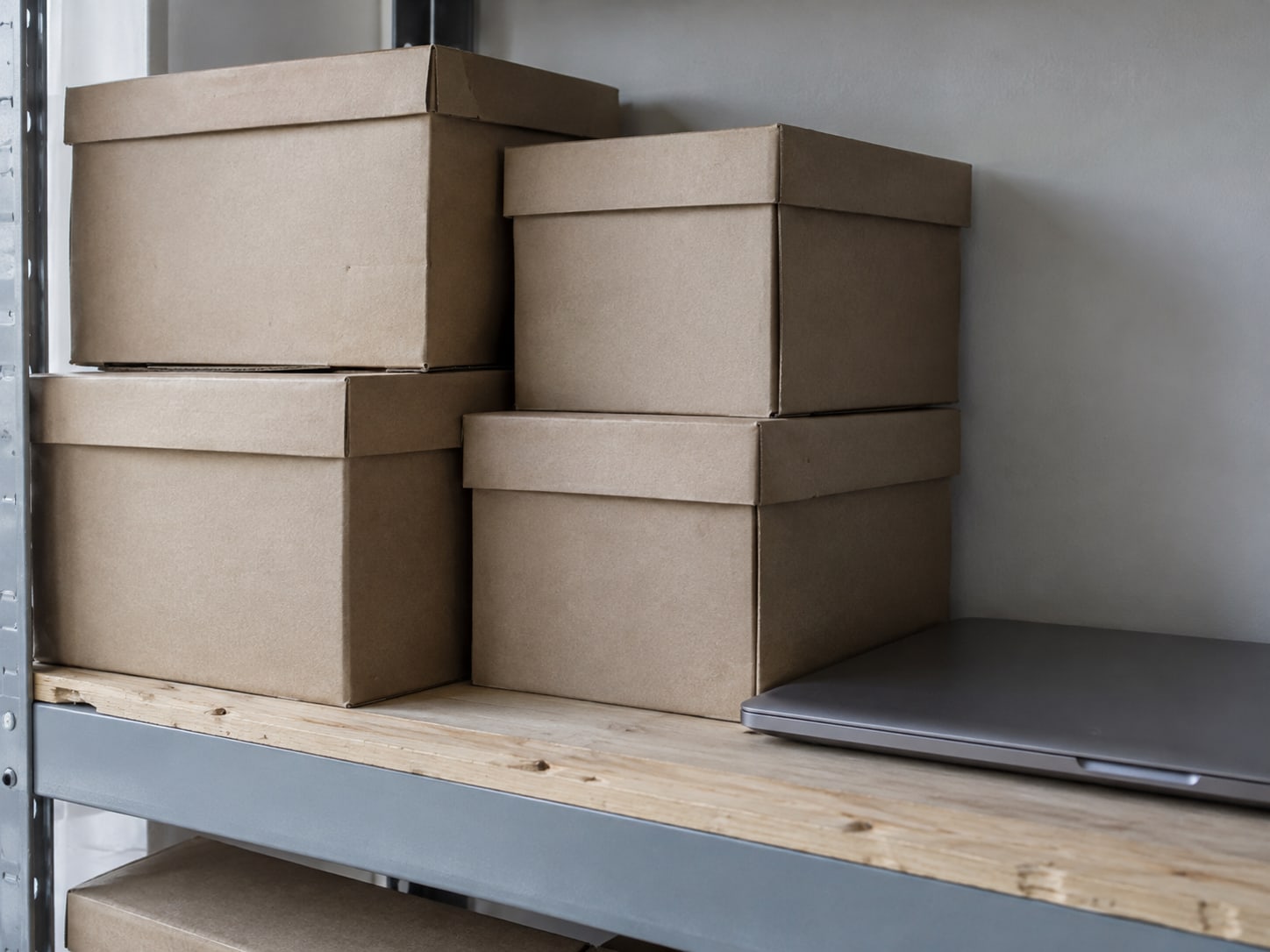 Several plain cardboard file boxes with blank labels stacked on a shelf beside a closed laptop.