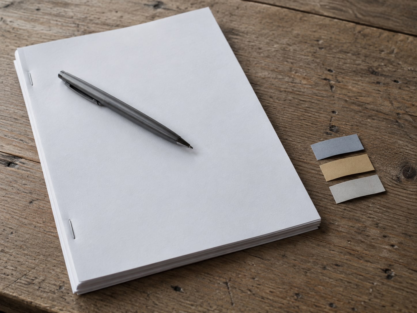 A pen resting on a stack of blank stapled pages beside three small colored sticky tabs on a wooden desk.