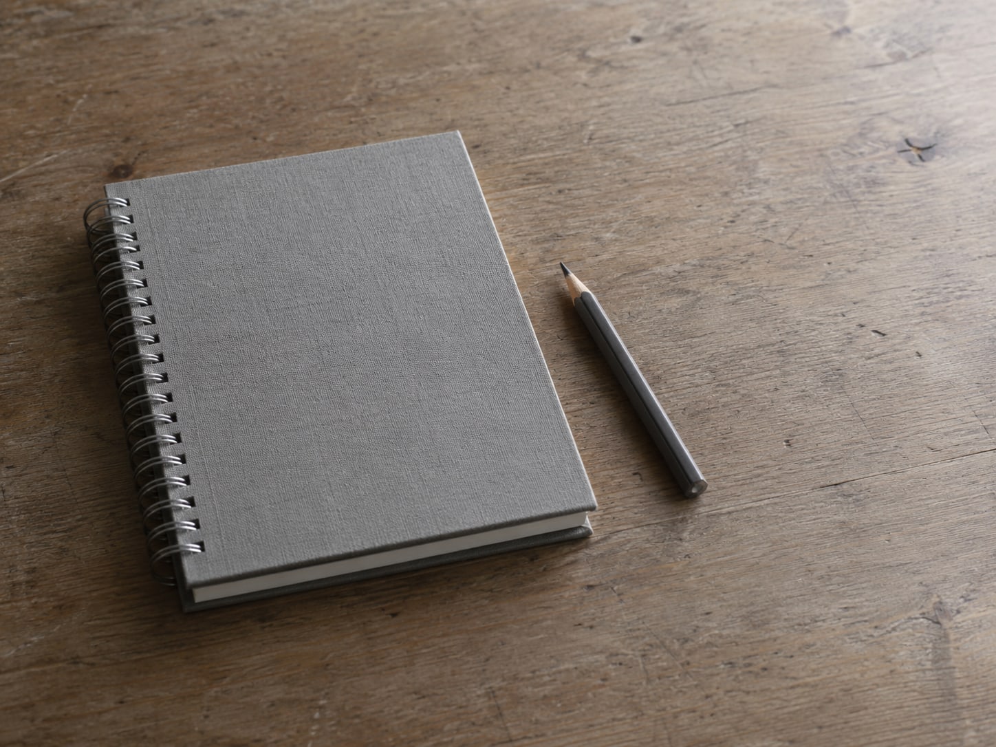 A closed spiral notebook and a short pencil resting on a bare wooden desk.