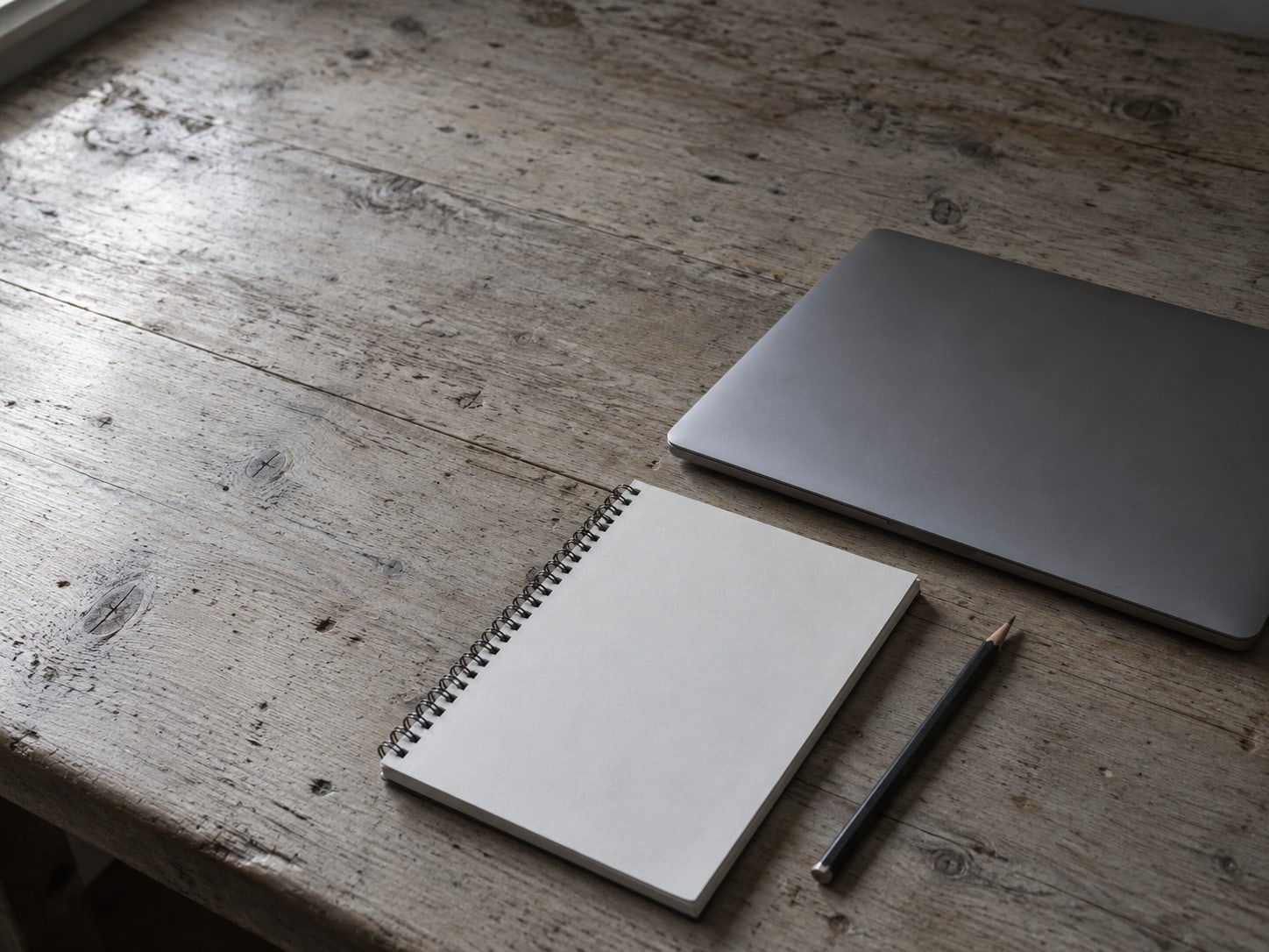 A closed laptop beside a blank notepad and a sharpened pencil on a worn wooden desk.