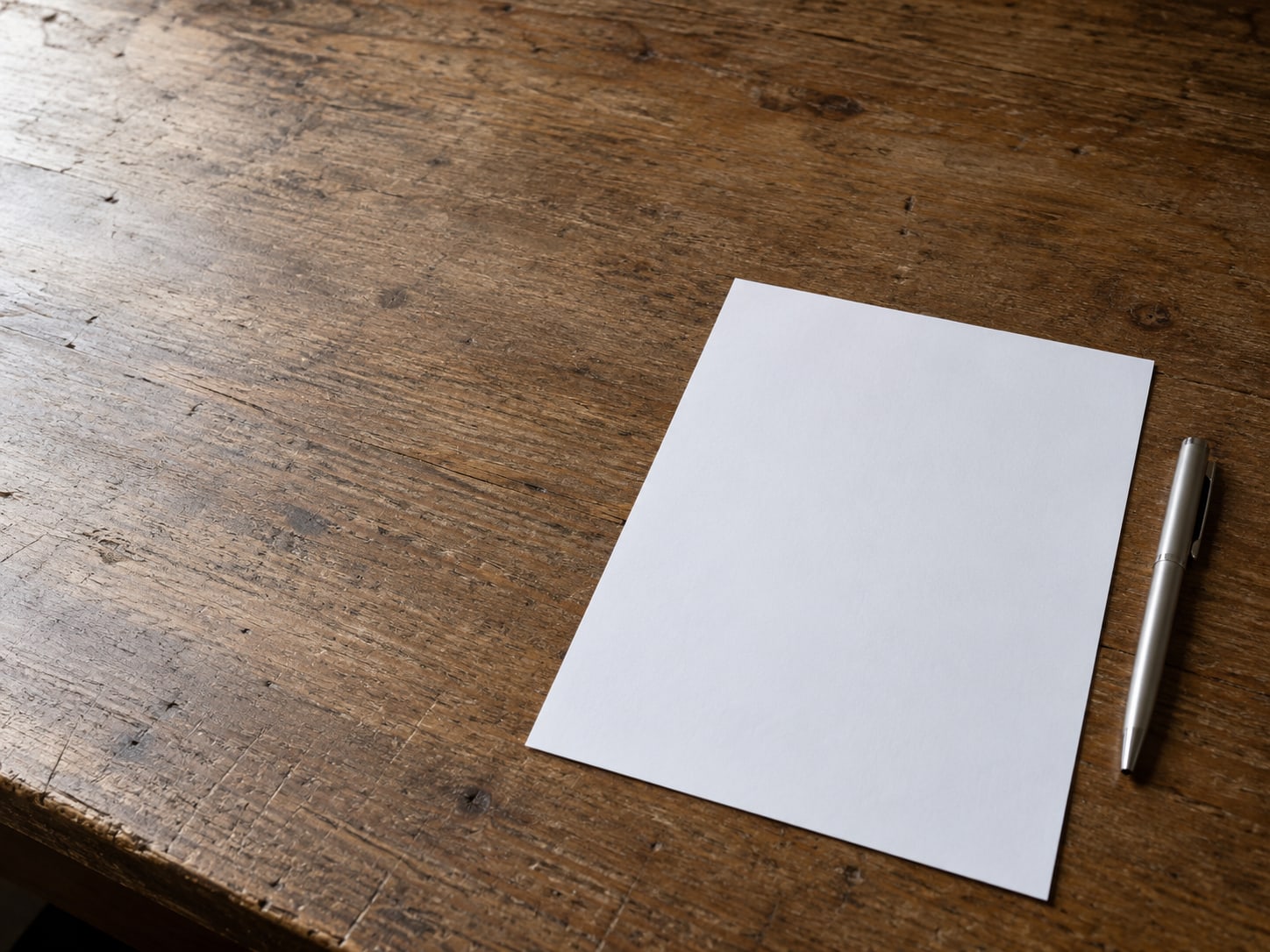 A single sheet of blank paper beside a closed pen on a plain wooden table.
