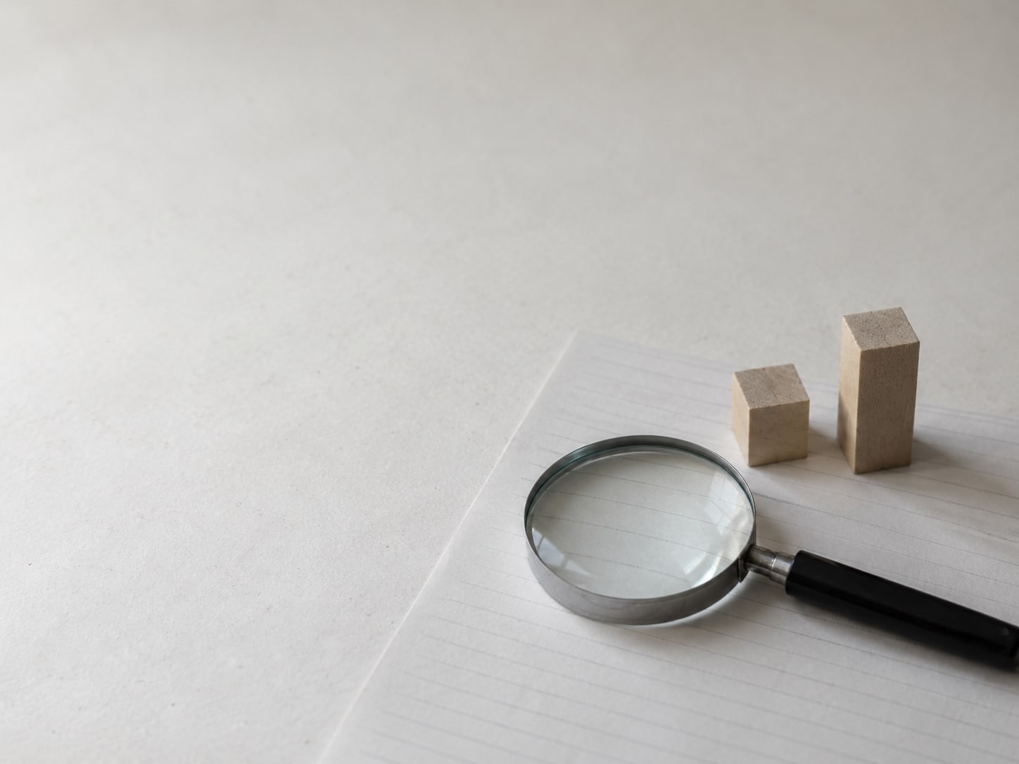 A magnifying glass resting on a blank sheet of lined paper beside two small wooden blocks, one taller than the other.