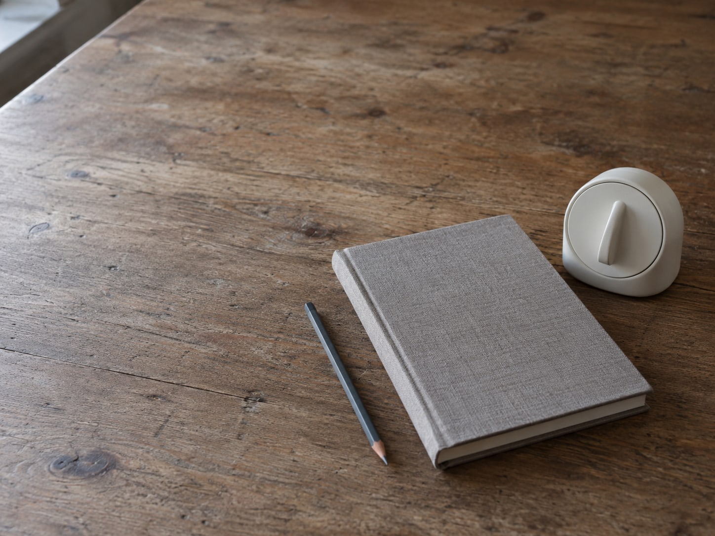 A closed notebook with a pencil beside a small kitchen timer on a plain wooden table.