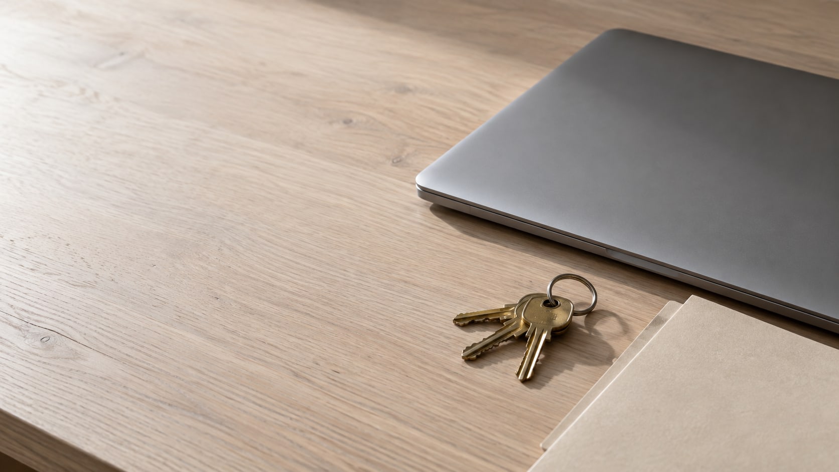 A closed laptop and a ring of brass keys on a pale oak desk beside a blank folder.
