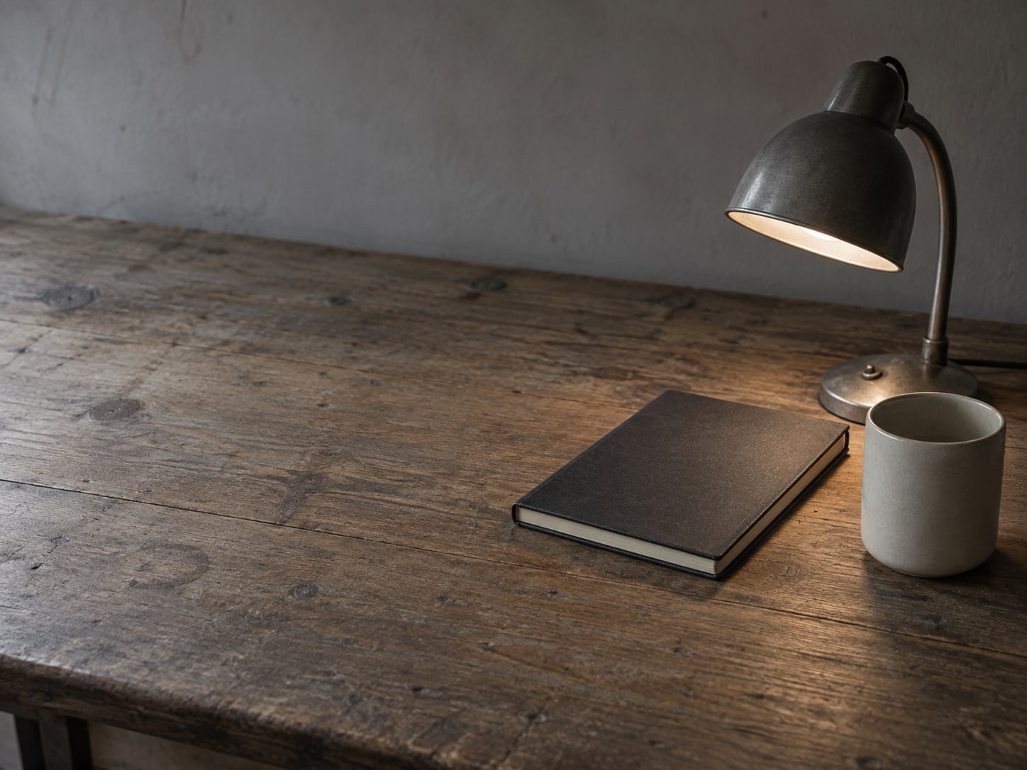 A single desk lamp lighting a wooden desk with a closed notebook and a mug.