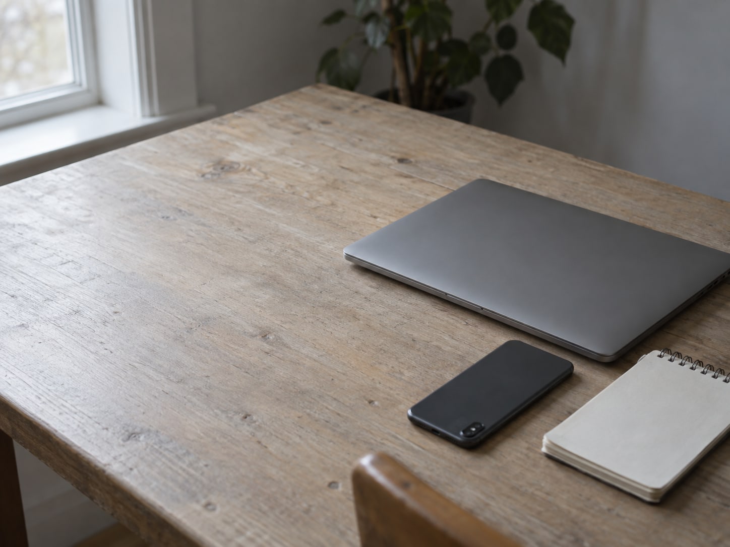 A closed laptop and a phone lying face down on a wooden desk beside a small blank notepad.