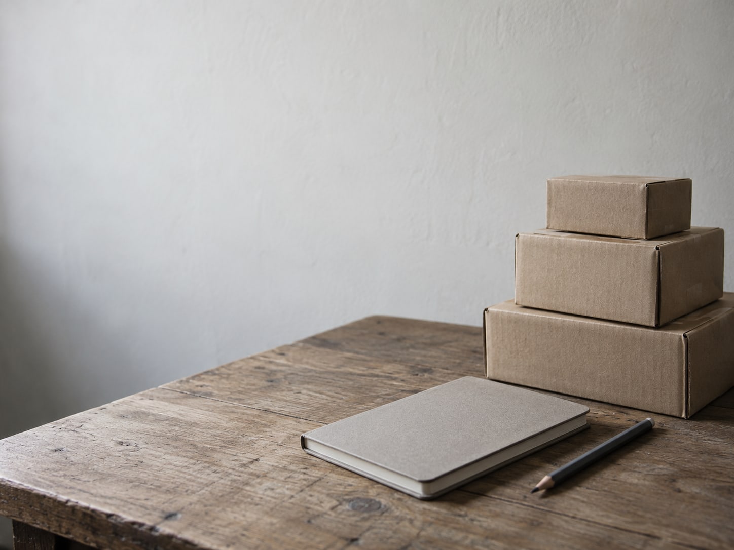 A closed notebook and a pencil beside a small stack of cardboard shipping boxes on a worn wooden table.
