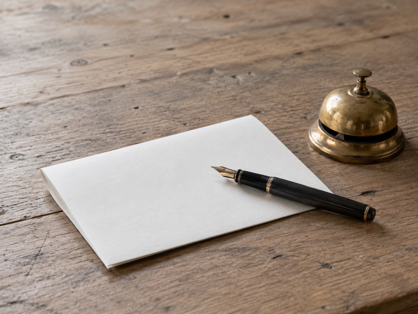 A fountain pen resting on a blank folded sheet of paper beside a small brass desk bell.