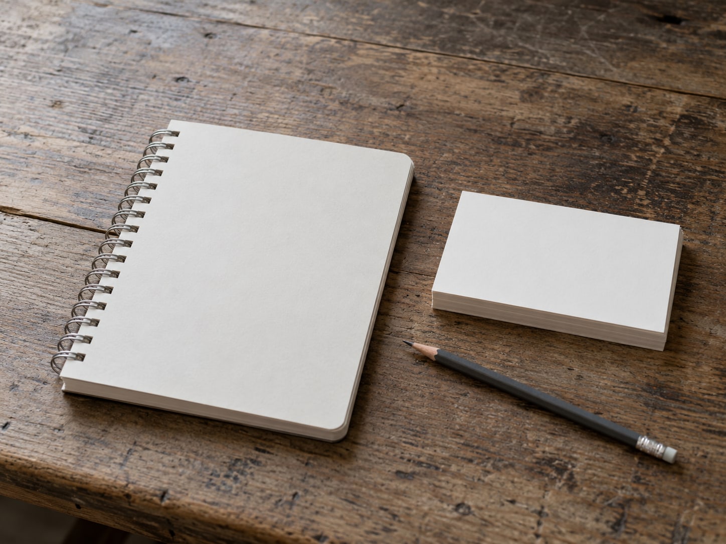 A closed spiral notebook and a pencil beside a small stack of blank index cards on a wooden table.