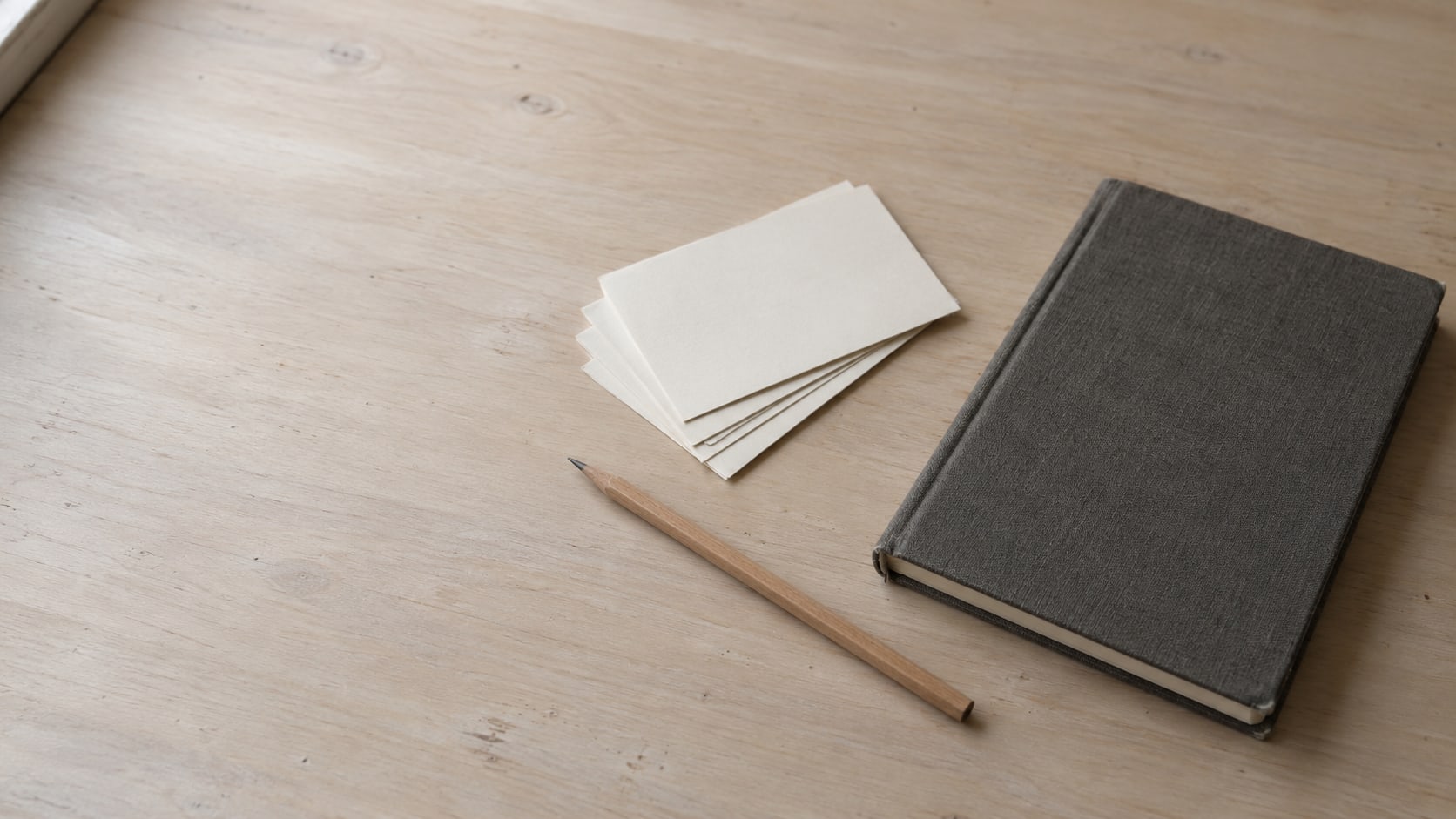 A closed notebook, a sharpened pencil and a fan of blank index cards on a plain wooden desk.