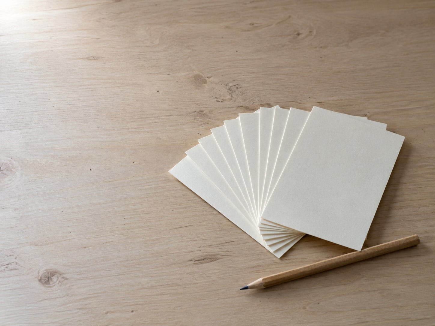 A fan of blank index cards beside a sharpened pencil on a plain wooden desk.