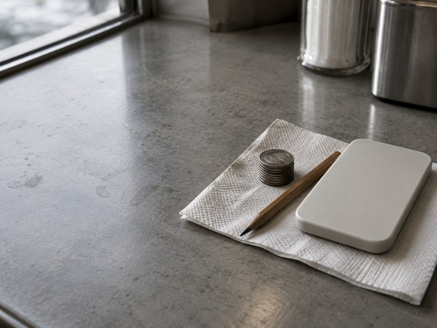 A small stack of coins, a pencil and a blank calculator sitting on a folded paper napkin on a diner counter.