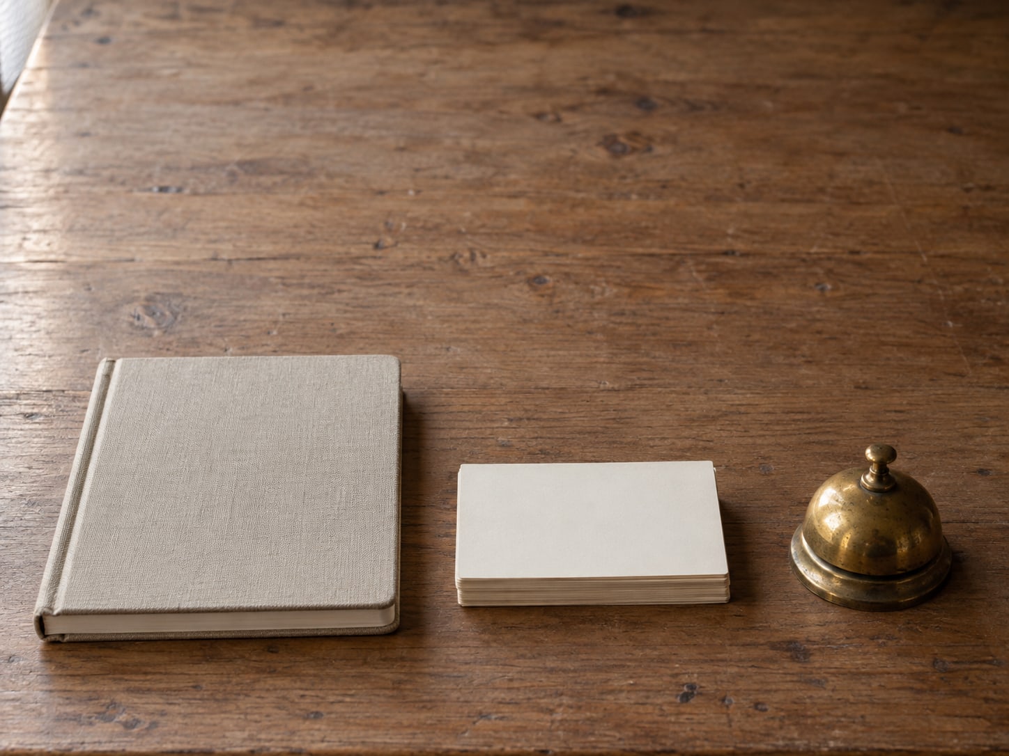 A wooden desk with a closed notebook, a stack of index cards and a small brass bell, arranged in a row from left to right.