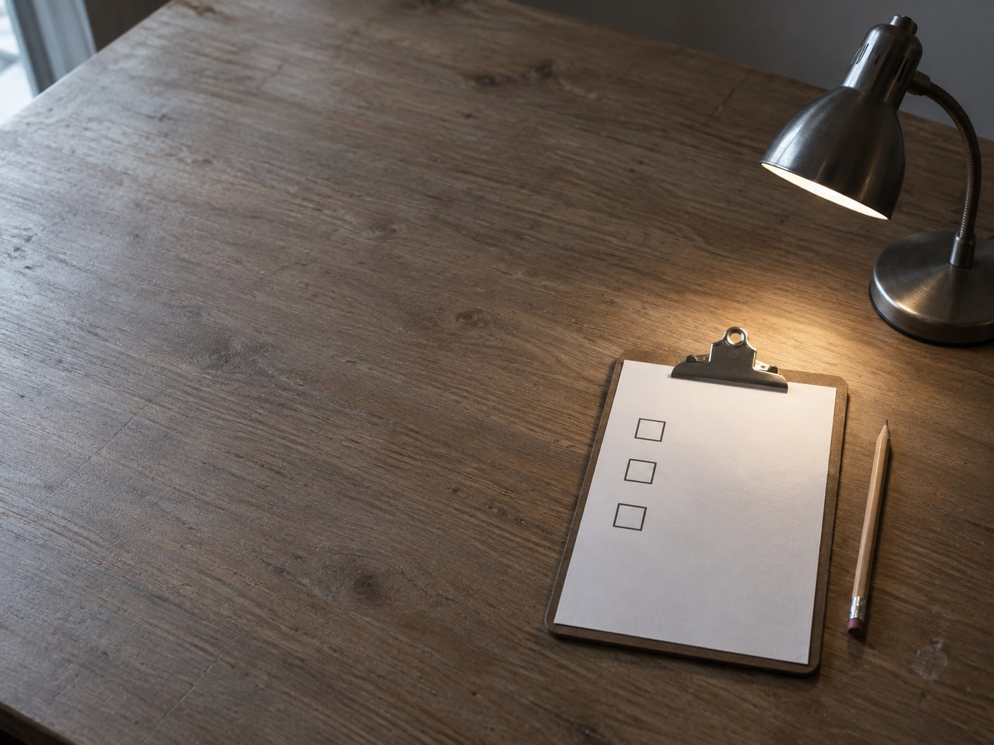 A small desk lamp shining on a plain clipboard with three blank checkboxes and a sharpened pencil beside it.
