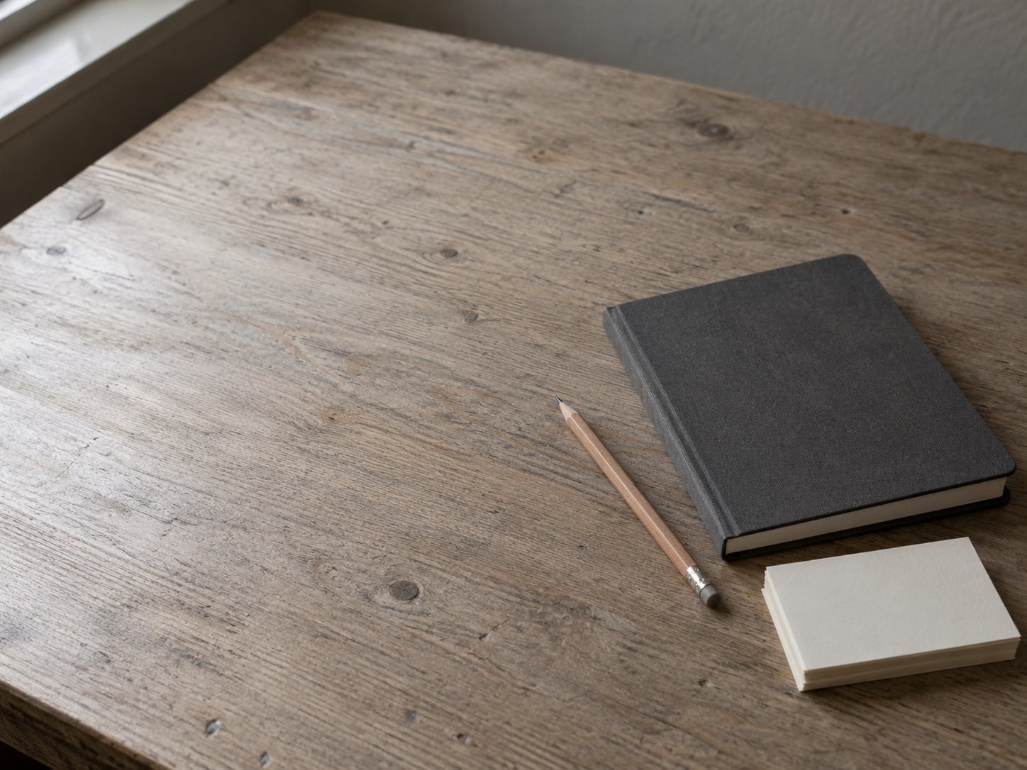 Un cuaderno cerrado junto a un lápiz y una pequeña pila de fichas en blanco sobre un escritorio de madera, listos para una lista de verificación.