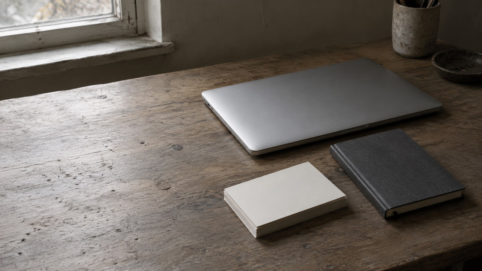 Una laptop cerrada, un cuaderno pequeño y una pila de tarjetas en blanco sobre un escritorio de madera.