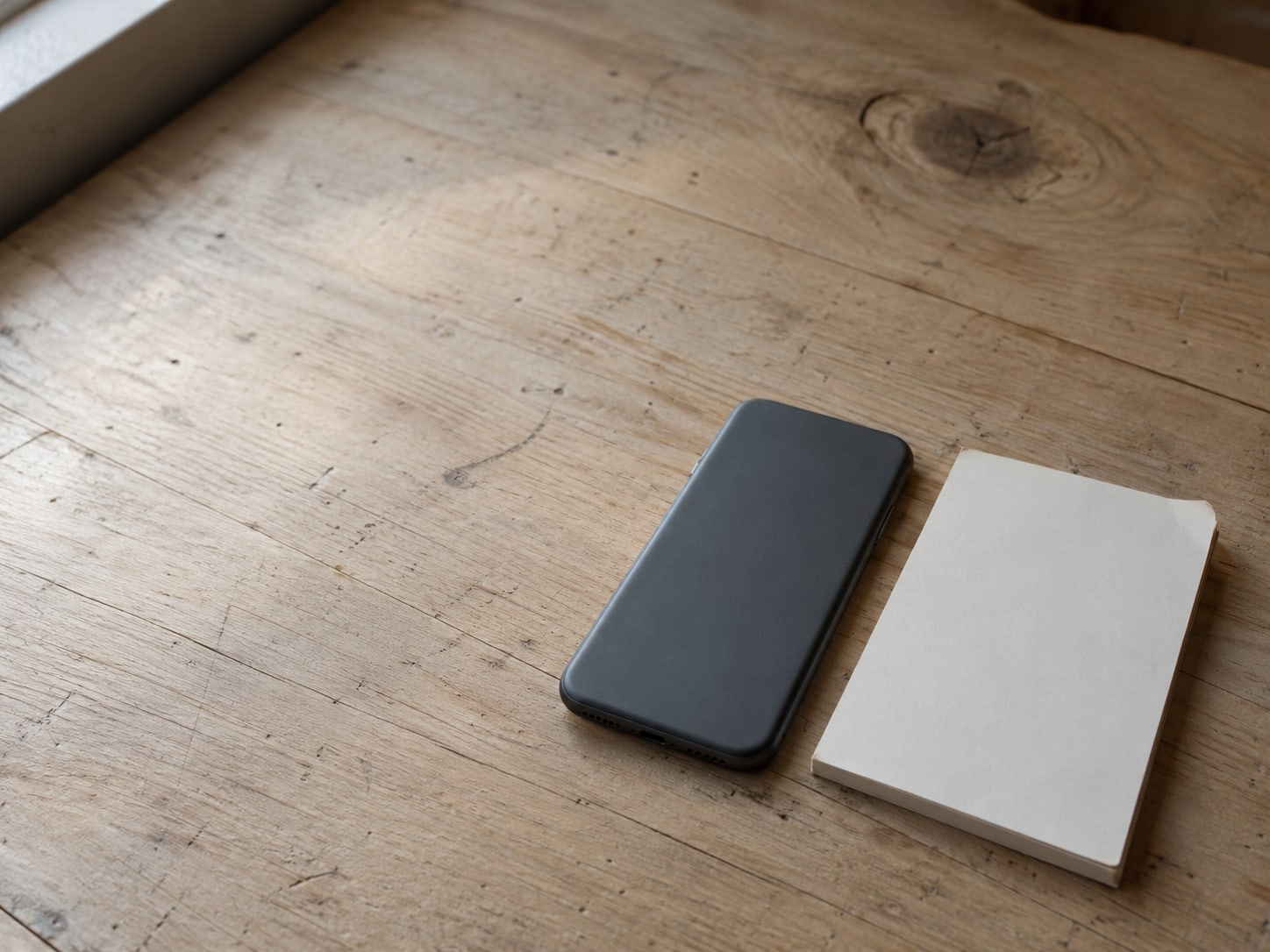 A smartphone lying face down on a wooden counter beside a small notepad with blank pages.