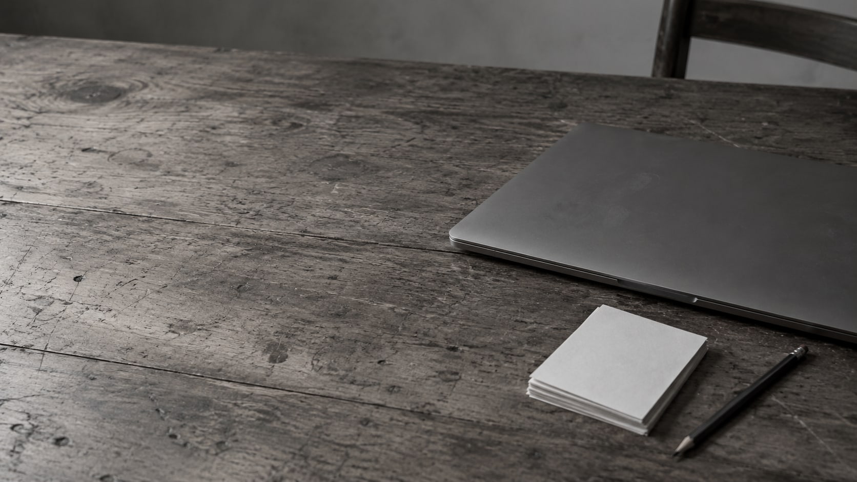 Una laptop cerrada junto a una pequeña pila de papelitos en blanco y un lápiz sobre un escritorio de madera desgastada.