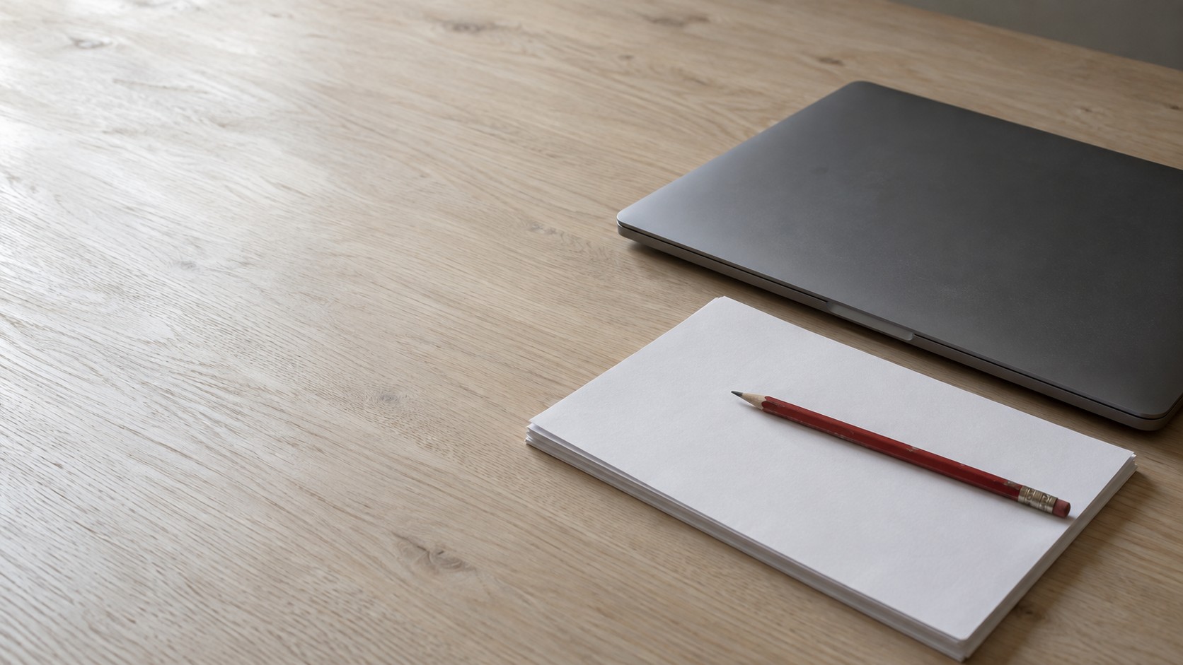 A closed laptop beside a stack of blank paper with a red pencil resting on top on a wooden desk.
