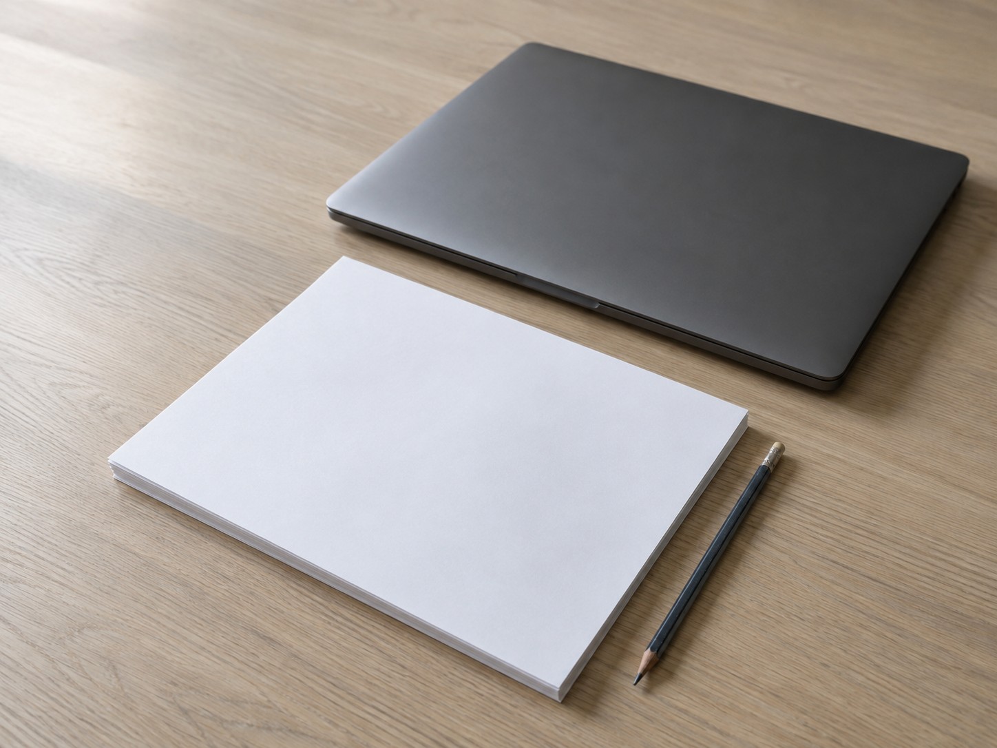 Una laptop cerrada junto a una pila ordenada de hojas en blanco impresas y un solo lápiz sobre un escritorio de roble claro.