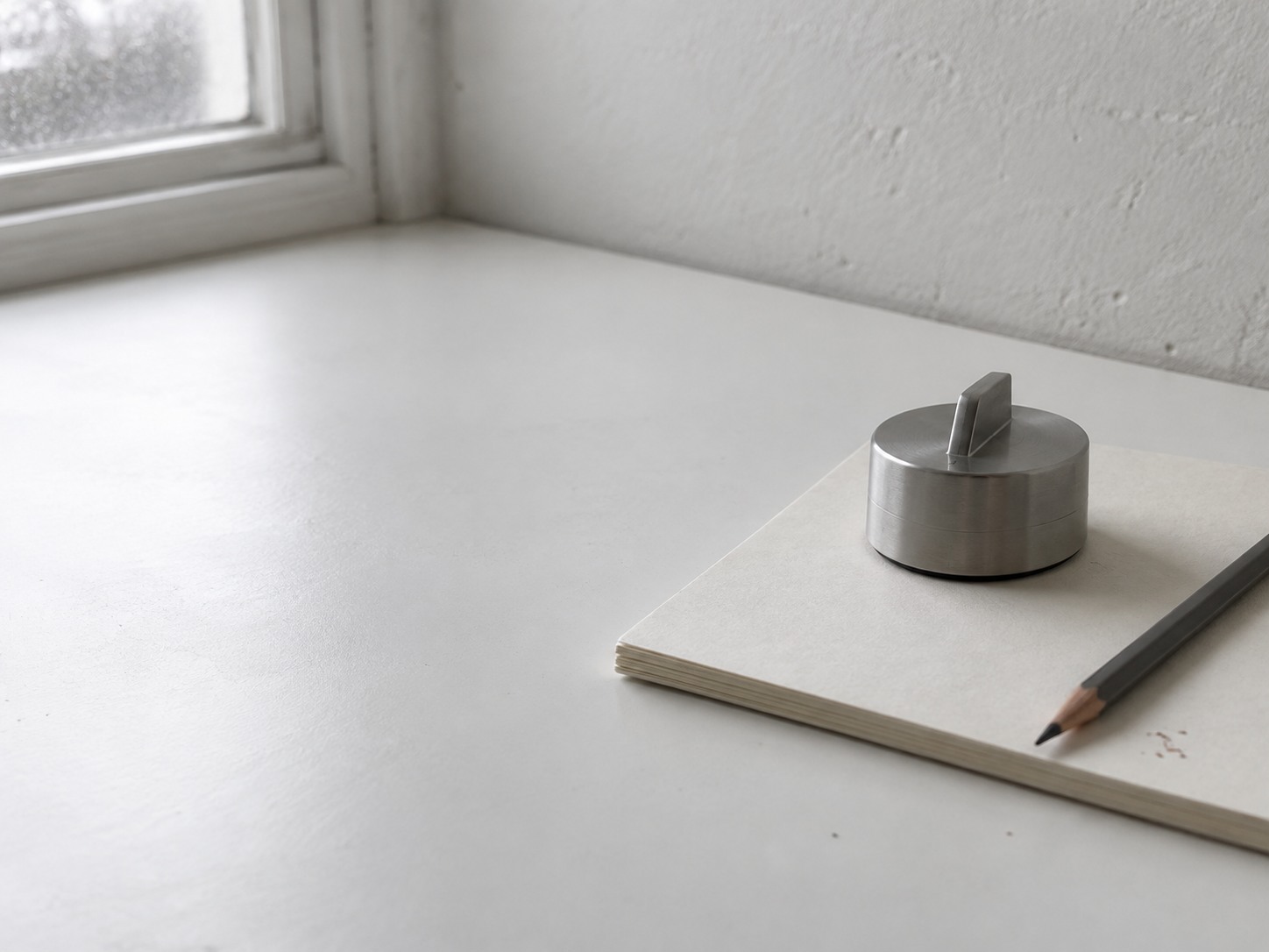 Un pequeño reloj de cocina descansando sobre una libreta en blanco junto a un lápiz afilado sobre un escritorio blanco.