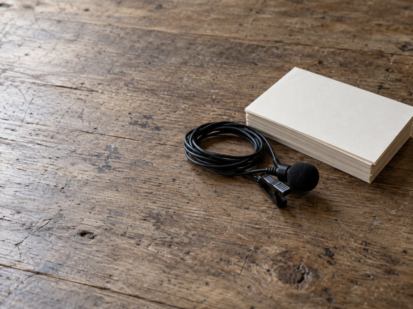A coiled lapel microphone beside a stack of blank index cards on a quiet wooden table.