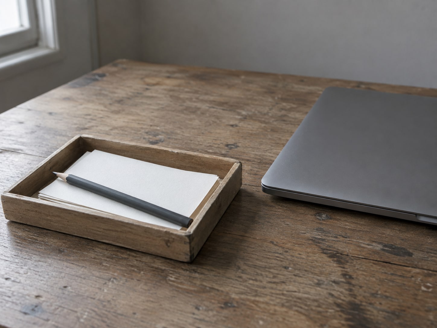 A small wooden tray holding a few blank index cards and a pencil beside a closed laptop on a desk.