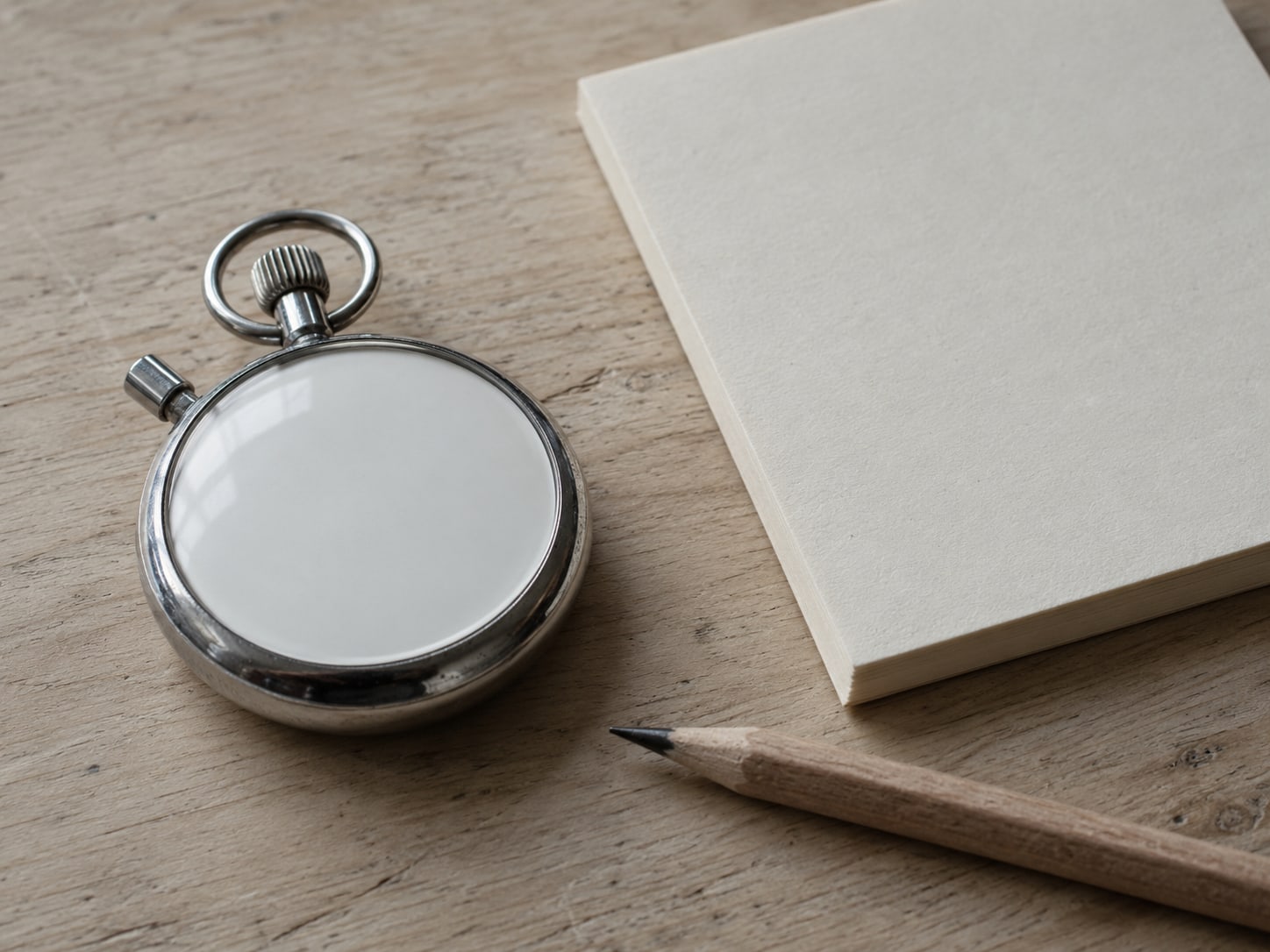 A stopwatch lying beside a blank notepad and a pencil on a light wooden desk.
