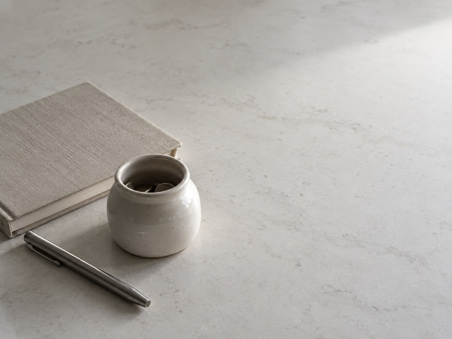 A small ceramic jar of loose coins beside a closed notebook and a pen on a clean countertop.