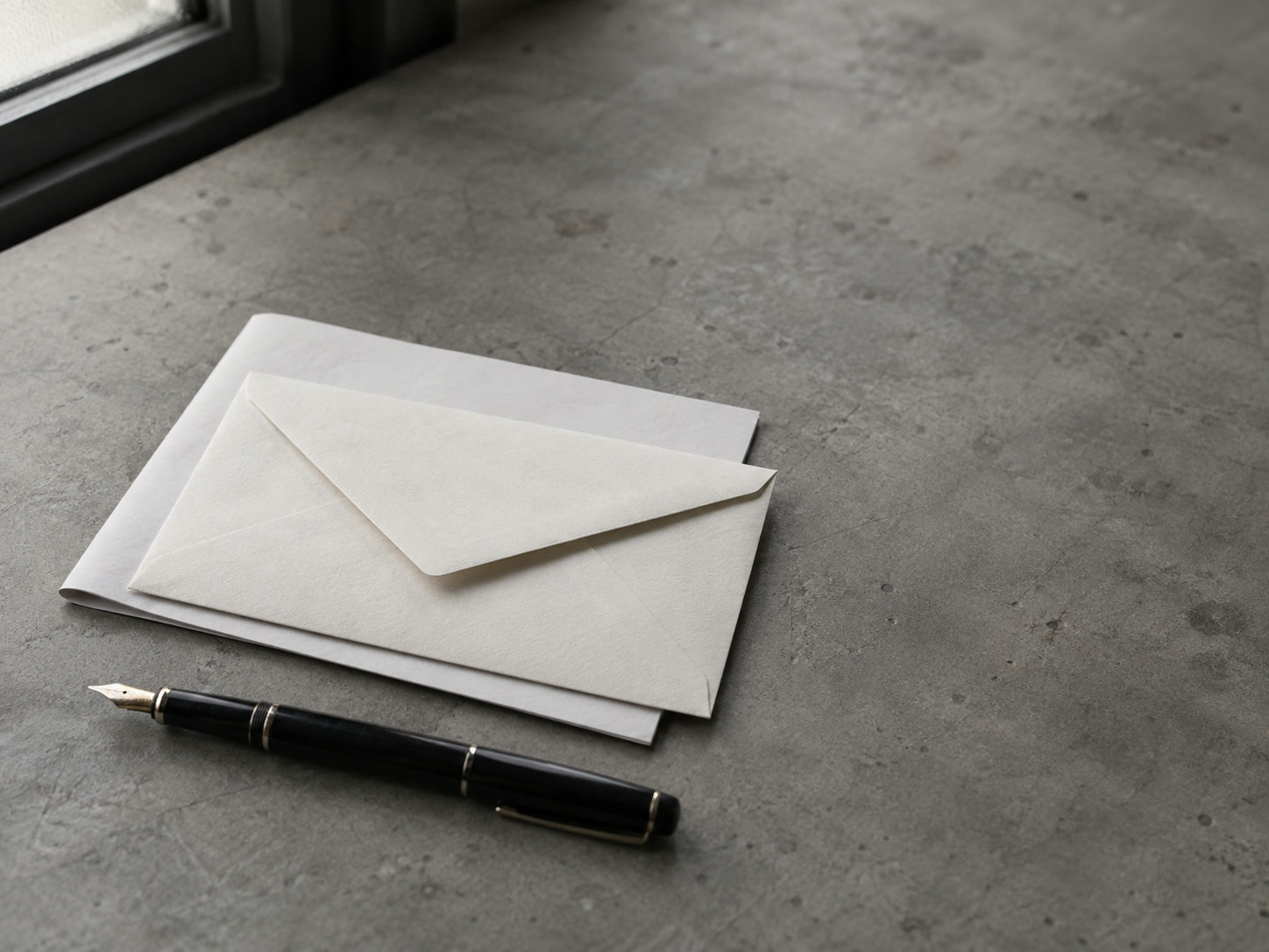 A sealed blank envelope resting on a folded sheet of plain paper beside a fountain pen on a gray desk.