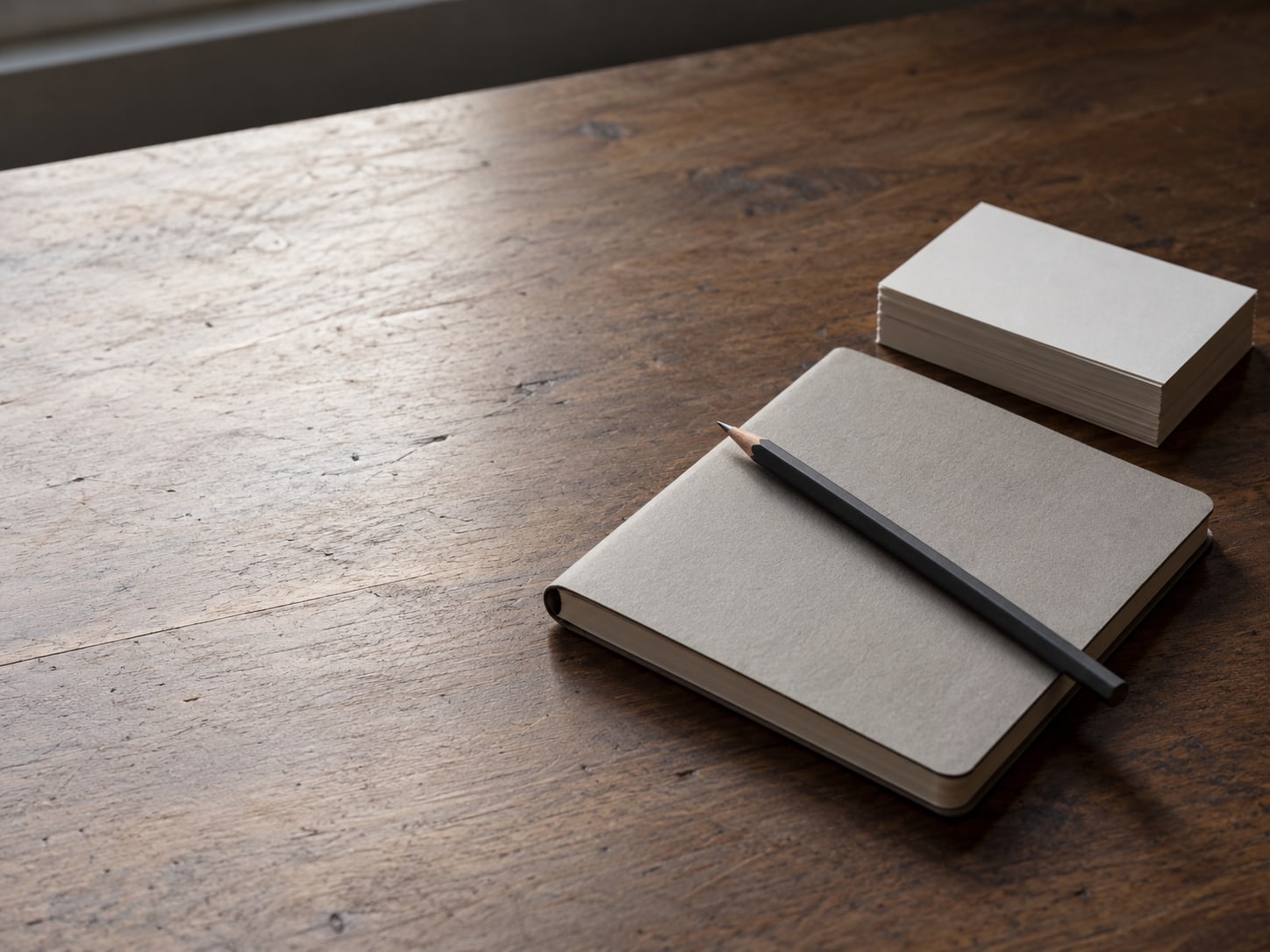 A closed notebook with a pencil resting on top, beside a small stack of blank index cards on a wooden desk.