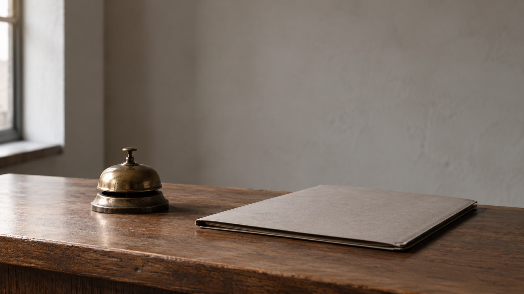 A small brass desk bell and a closed paper folder resting on a clean wooden counter in soft window light.