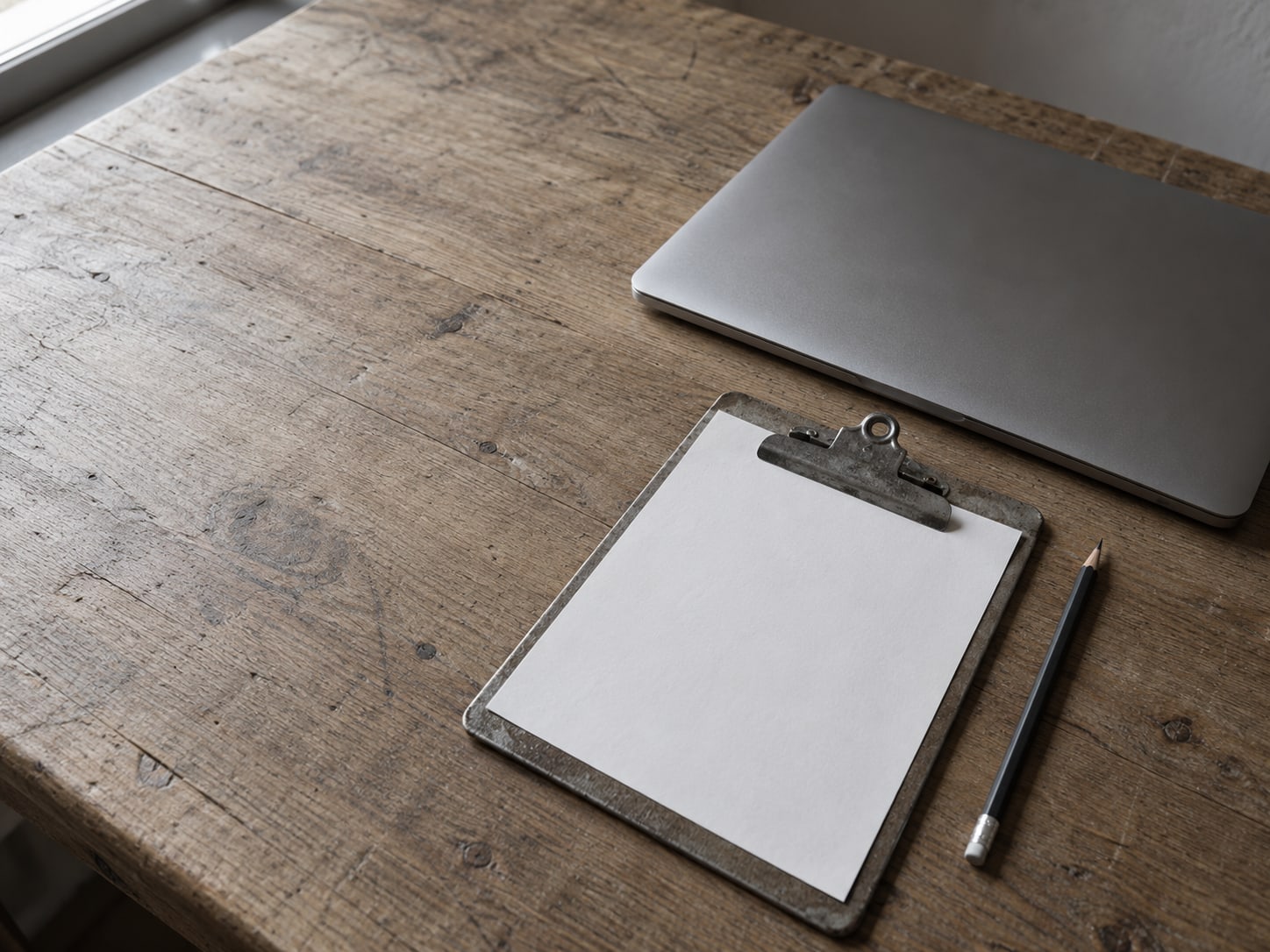 A clipboard with a blank checklist and a pencil lying beside a closed laptop on a plain desk.