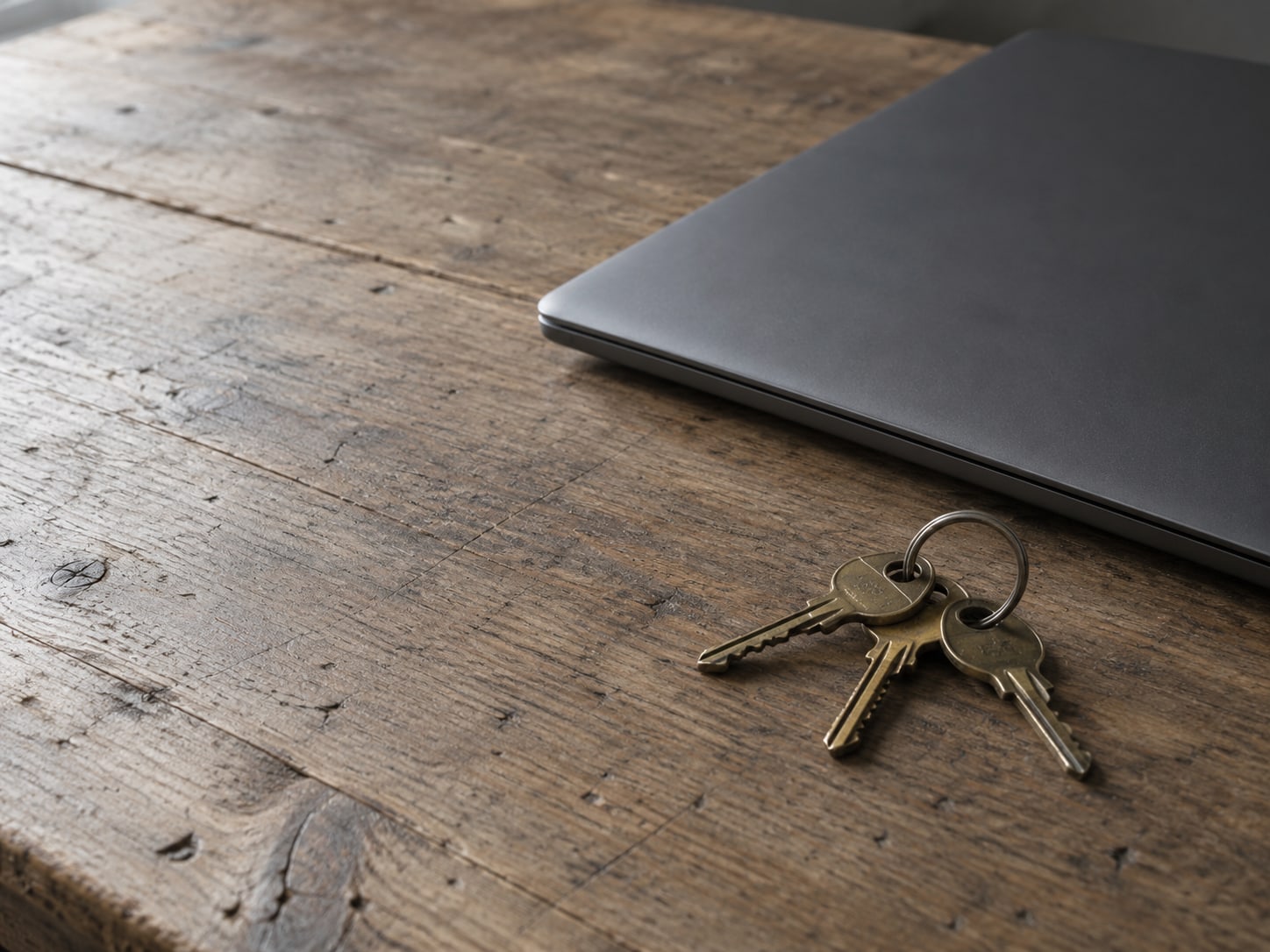 A closed laptop beside three small brass keys on a plain ring, resting on a worn wooden desk.