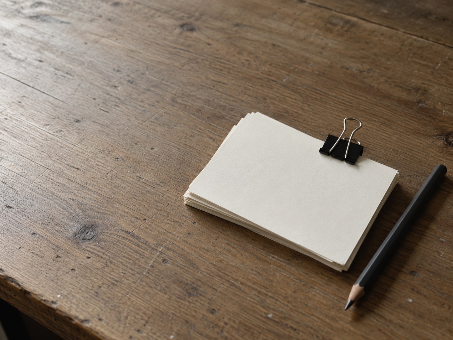 A small stack of blank index cards held with a binder clip beside a plain pencil on a wooden desk.