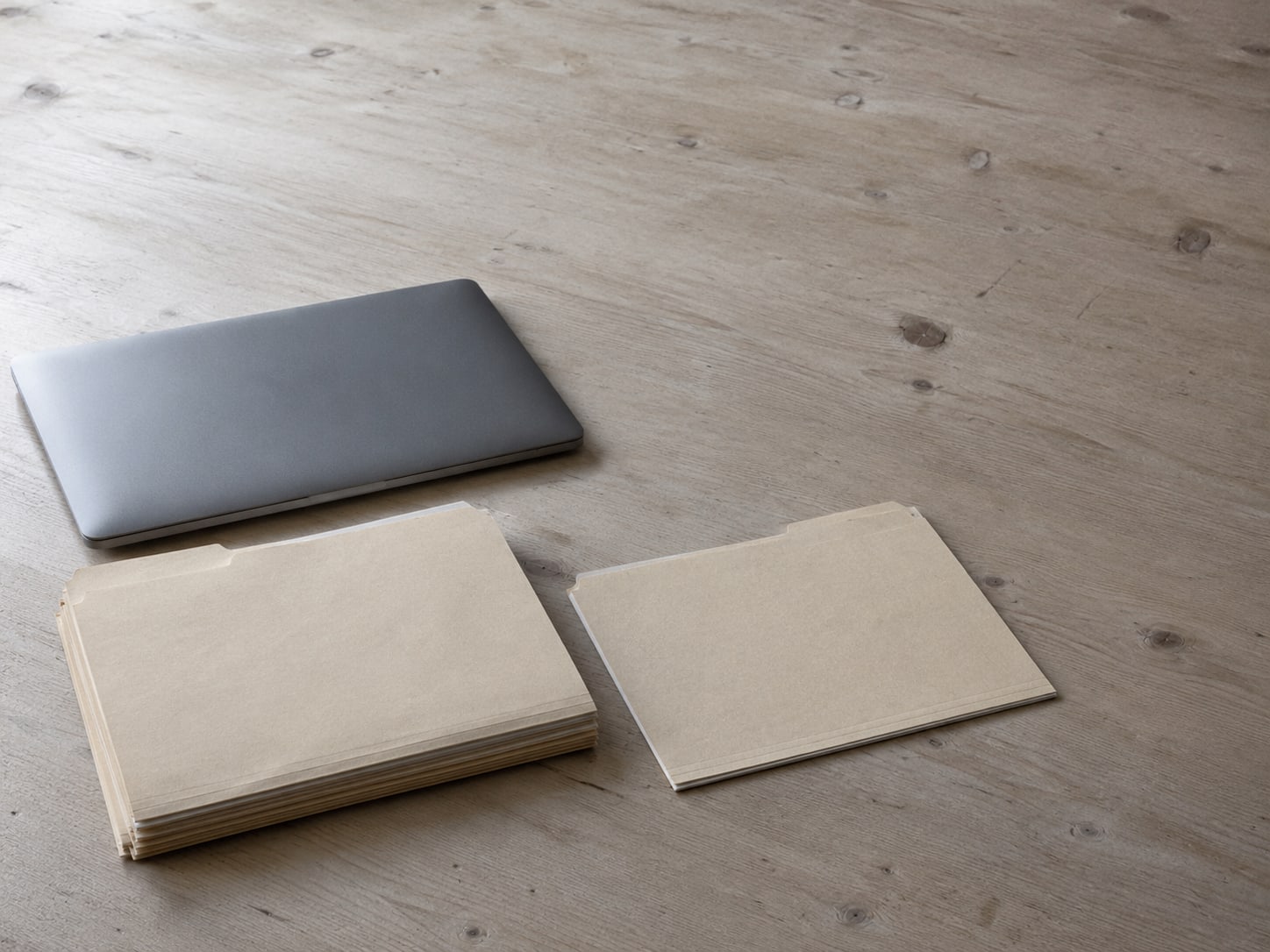 A closed laptop beside two manila folders, one thick and one thin, on a plain desk.