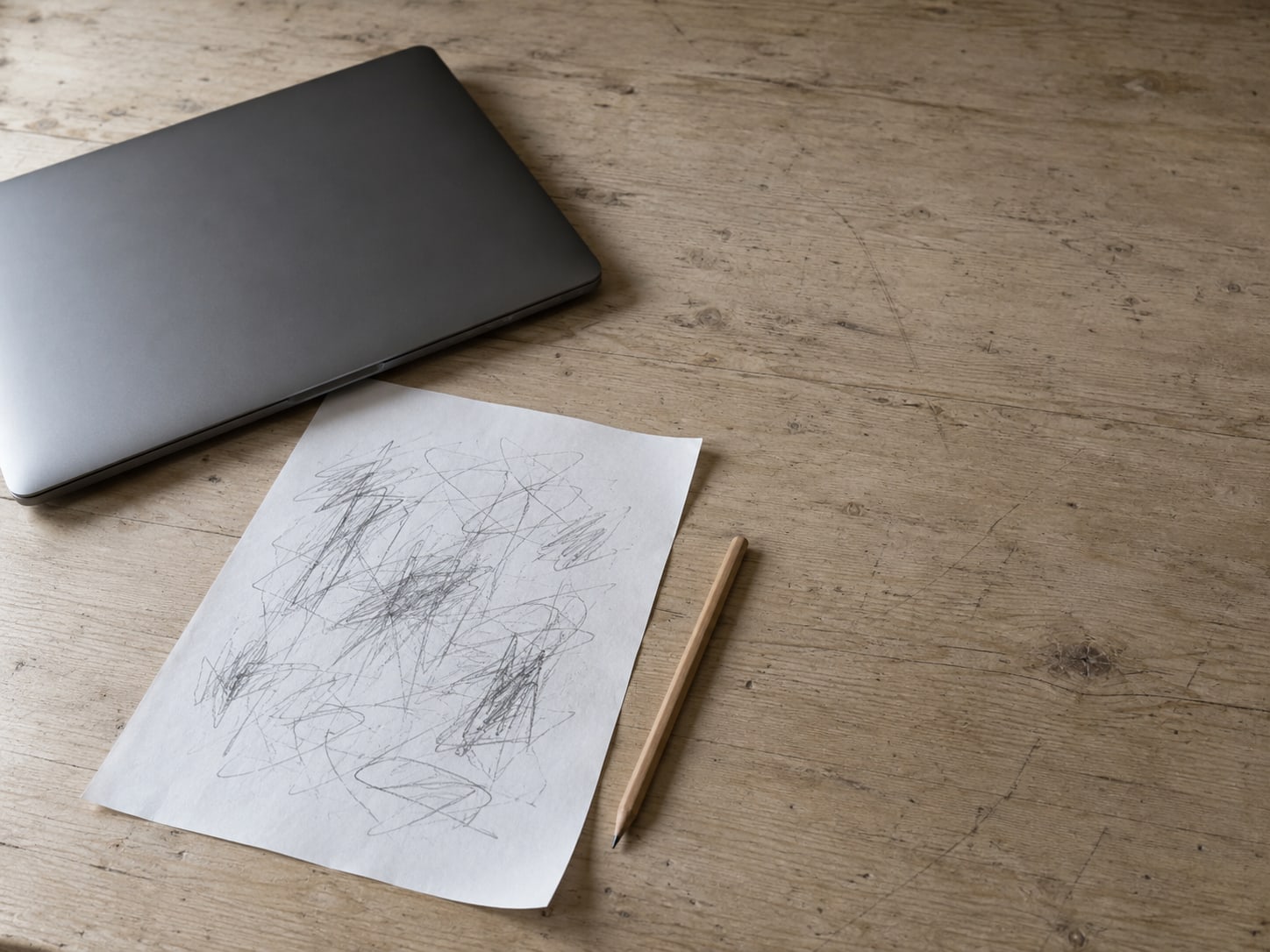 A printed page lying on a wooden desk, covered in unreadable pencil marks, with a pencil and a closed laptop beside it.