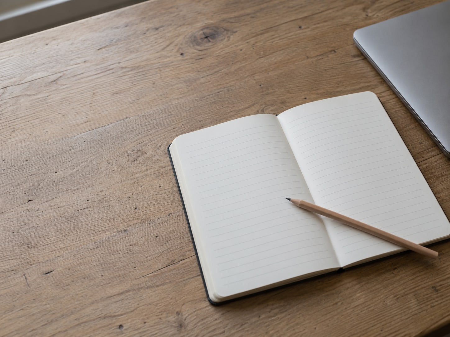 An open notebook with two columns of blank lines and a pencil resting across the page, beside a closed laptop on a desk.