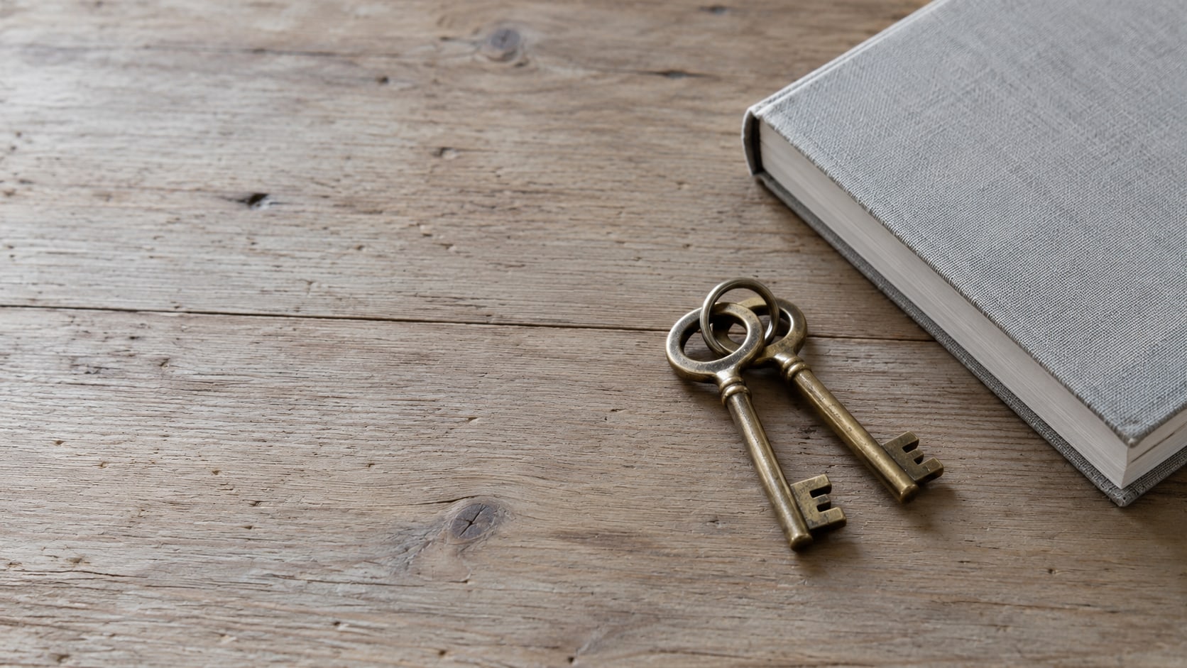A pair of identical brass keys resting on a bare wooden tabletop beside a closed notebook.