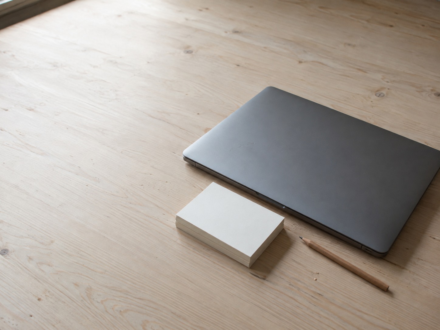 A closed laptop beside a neat stack of blank index cards and a pencil on a pale wooden desk.
