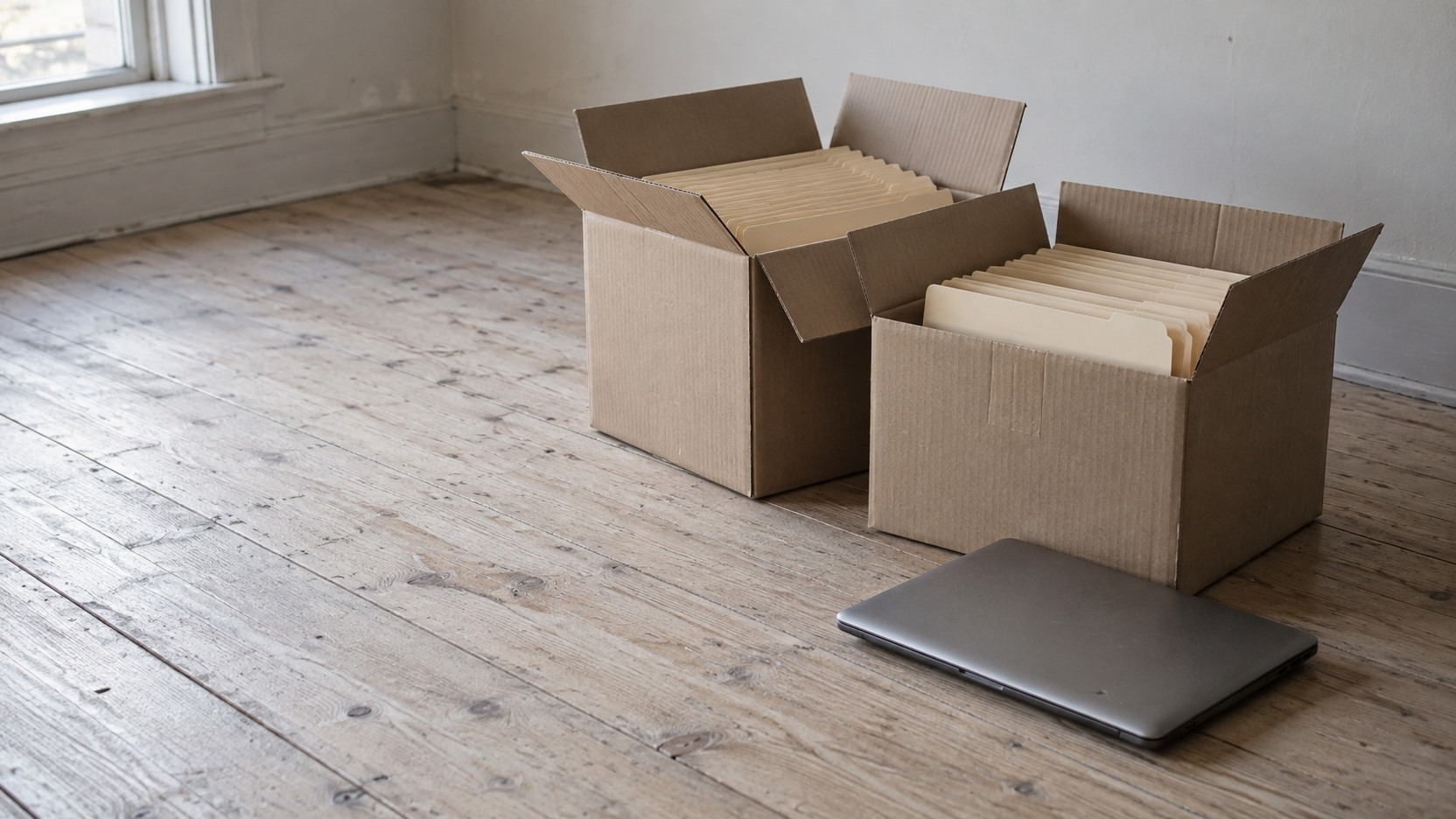Two open cardboard boxes of blank folders beside a closed laptop on a wooden floor.