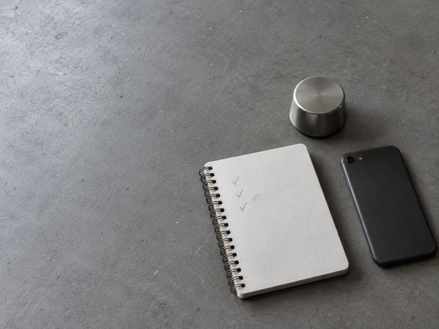 A spiral notebook with faint pencil tick marks beside a closed phone lying face down and a small desk timer on a grey table.