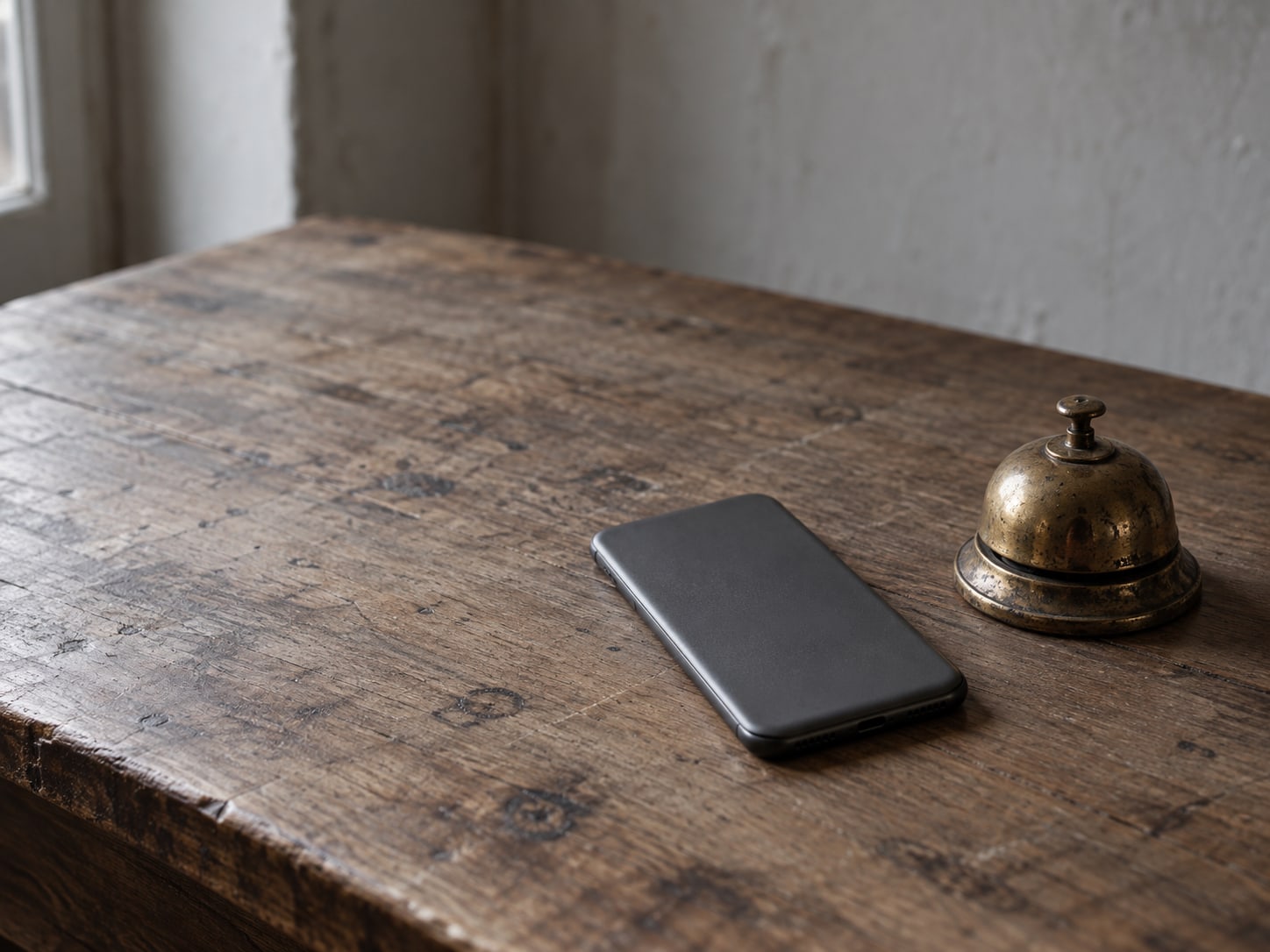 A smartphone lying face down on a worn desk next to a small brass bell.